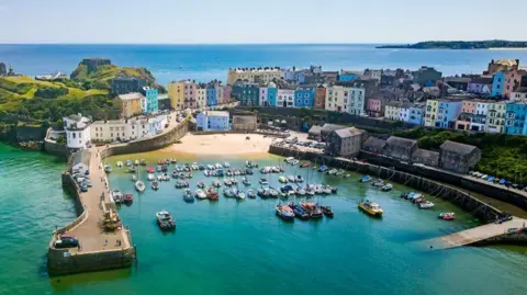 Getty Images About 80 small boats moored in green-blue water in the harbour in Tenby, viewed from above in bright sunshine. There are rows of multi-coloured houses behind the harbour - green, blue, yellow, turquoise, orange and pink - and a view out to sea at the top of the frame.