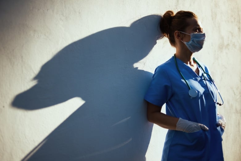 A tired-looking female health-care worker wearing medical scrubs, a medical mask and rubber gloves, with a stethoscope around her neck, leans against an outdoor wall in the sunshine, which casts her shadow.