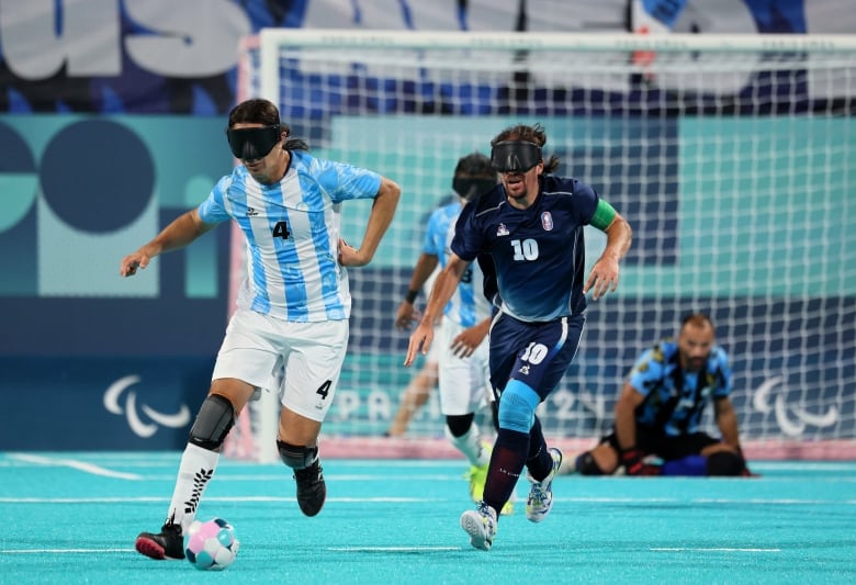 Two men on different teams run towards a soccer ball during a game while wearing dark blinds over their eyes. 