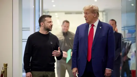 Shutterstock Ukrainian President Volodymyr Zelensky, dressed in a black sweater with a Ukrainian coat of arms emblem, engages in conversation with President-elect Donald Trump, wearing a blue suit, white shirt, and bright red tie.