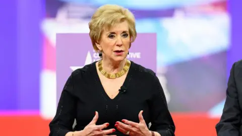 Getty Images Linda McMahon, who has short blonde hair and is wearing a black dress with a thick golden necklace, gestures with both hands in her seat during a Conservative Political Action Conference (CPAC) panel