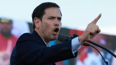 Getty Images Marco Rubio, wearing a suit, gestures with the index finger of his right hand, as he speaks at a Trump rally