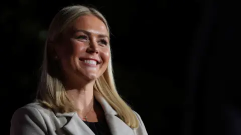 Reuters Karoline Leavitt, who has straight blonde shoulder-length hair, and wears a silver cross necklace with a cream coat, beams during a rally in 2022