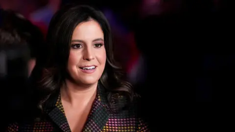 Getty Images Elise Stefanik, with her black shoulder-length hair down and wearing a multi-coloured suit, speaks with members of the media