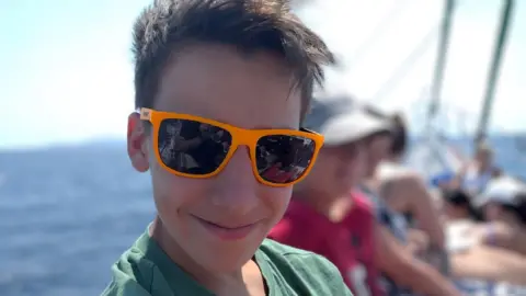 Murray looking at the camera and smiling while wearing bright orange sunglasses. He appears to be on a boat, with the sea visible behind him and other people sitting alongside him.