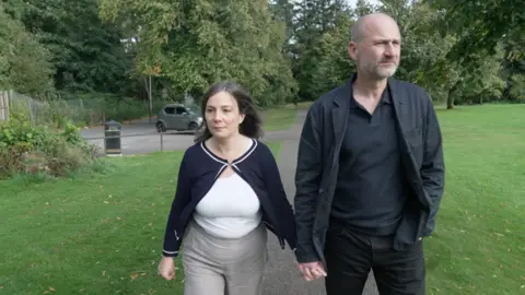 Ros and Mark holding hands as they walk in a park. Mark is wearing a black shirt and jacket. Ros has a white top on with a black jacket.