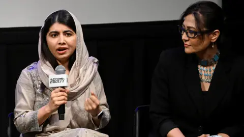 Getty Images Malala Yousafzai and Sahra Mani on stage at an event in New York. Malala holds a microphone as she addresses the audience. She wears a pale grey dress and headscarf, embellished with gems. Sahra sits to her left and looks at her as she speaks. Sahra wears a black top and blazer with an elaborate blue necklace. 