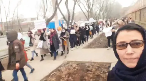 Apple TV+ A still from Bread & Roses of a women's march. A long line of women walk along a path in Kabul holding placards while a woman dressed in a black dress and headscarf in the right-hand corner of the image takes a selfie. The city seems wintry, with no leaves on the trees and many of the protesters dressed in warm coats. 