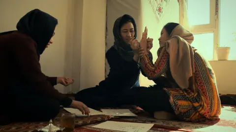 Apple TV+ A still from Bread & Roses showing three women sitting together on the floor. The woman in the middle writes on the hand of the woman sitting to her left while the third woman looks at sheets of paper spread on the carpet. 