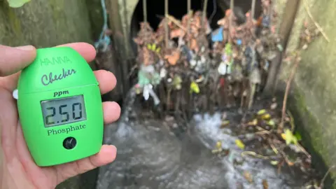 CURB A hand holding a green phosphate reader in front of sewage outfall. The device shows a level of "2.50ppm".