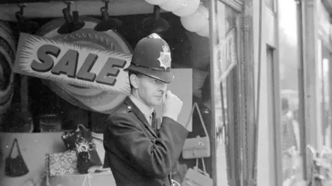 Getty Images A uniformed police officer stands outside the SAC shop. He is wearing a tall helmet and is scratching his nose with his thumb