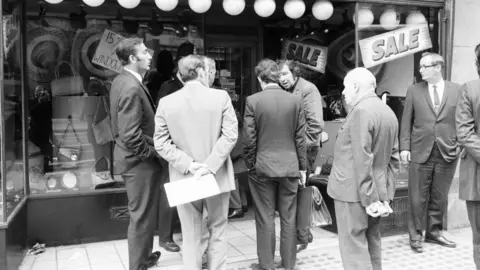 Getty Images Detectives in suits gather outside SAC, a leather goods shop on Baker Street. They are all looking at each other in a cross manner