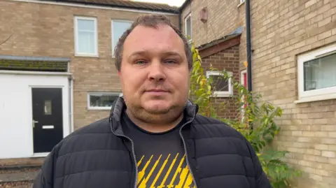 Kate Bradbrook/BBC A man in a unzipped black coat. He is wearing a black t-shirt with a yellow striped design. He is standing outside a block of flats.