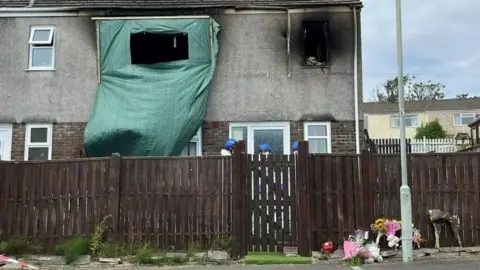 Fire damage can be seen on the exterior of the family's house, with the two windows on the first floor missing and smoke damage around the smaller of the two windows. Three people wearing white outfits and blue helmets are talking outside the front door. A number of floral tributes and teddy bears can be seen on the pavement outside.