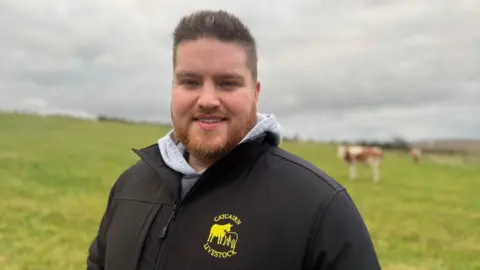 A man, standing in front of a field of grass, looking at the camera. He is pictured from the chest up. He is wearing a grey hoodie and a black jacket with a yellow logo with a cow on it. He has short brown hair and a tight beard. There is a cow in the background to the right of the man. The sky is full of grey clouds. 