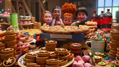 Netflix Children looking delighted as they look over a table filled with pizza, fizzy drinks, doughnuts, mince pies and other Christmas food, in a promotional still from Netflix's That Christmas