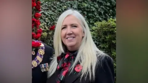 Janice Kirby Headshot of blonde woman in a black jacket, wearing a poppy 