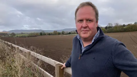 BBC/Carla Fowler A man, wearing a navy quarter zip fleece and check shirt, leans against an outdoor wooden fence. The background is of a muddy ploughed field. 