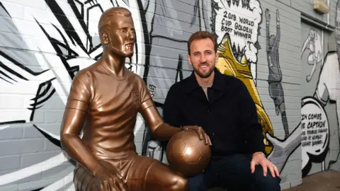 PA Media Harry Kane is smiling and sitting to the right of the life-size statue of himself, which is also in sitting position with a football perched on its knee. The statue is wearing the England kit. There is a black, white and grey mural behind.