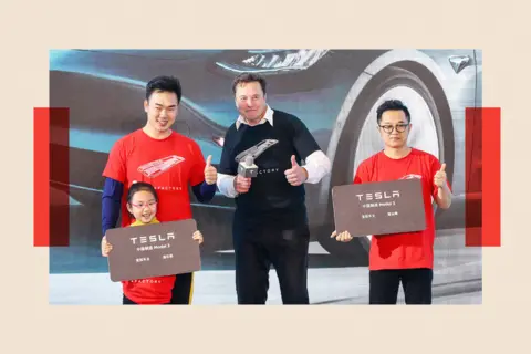 Getty Images Elon Musk poses for photos with his thumbs up. He is in between buyers who are holding Tesla signs, also with thumbs up.