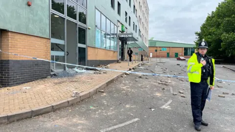 Smashed glass from a hotel lies on the pavement and rubble in the road as a member of the police in a neon jacket a police hat stands to the right of the photo in front of a cordon, holding a walkie talkie to his chest with his right hand and papers in his left hand.