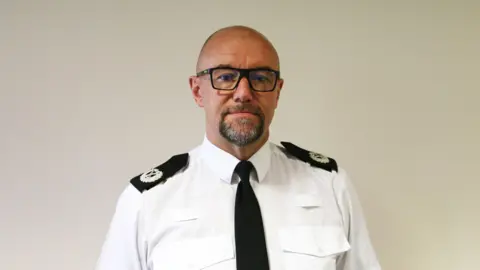 Staffordshire Police Man with square black glasses, grey beard and smooth head in a white police shirt with crests on his shoulders. His expression is neutral as he looks into the camera.