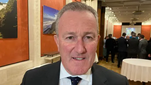 Conor Murphy - a man with short, grey hair smiles at the camera. He is standing in a large hall with orange walls. He is wearing a dark suit jacket, a white collared shirt and dark tie.