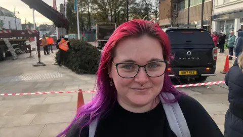 John Devine/BBC Emily Parkes has long red and purple hair over her shoulders. Her rucksack straps are visible. She has a piercing under her nose and is wearing spectacles. Behind her is a taped-off area where workmen are shaping the bottom of the trunk of the Christmas tree, which is lying on the pavement