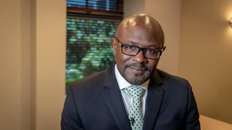 Bald man with a black beard wearing glasses, navy suit and light blue spotted tie.