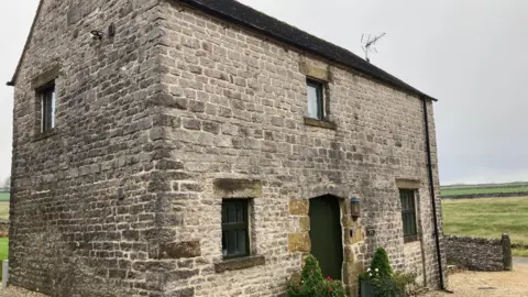 The picture shows a small stone-built cottage, which is used as a self-catering holiday property. It's in the tiny Derbyshire hamlet of Hurdlow. You can see the Peak District countryside in the background