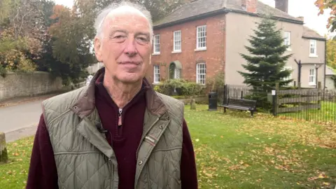 The picture shows Derbyshire Dales Councillor Peter O'Brien. He is standing on a grassed area in the Derbyshire village of Eyam