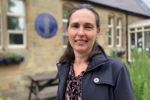 The picture shows the headteacher of Eyam Church of England Primary School, Oona Gilbertson. She is wearing a black jacket and is standing outide the front of the school