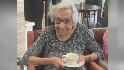 Handout Betty sits in a red chair while holding a cup of tea and smiling as she looks at the camera. She is wearing a grey patterned top and there is a round table behind her.