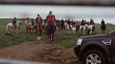 Mr John sits on top of his horse in a red hunting jacket. There are men on horses behind him in the field and a 4x4 to the right of the image.