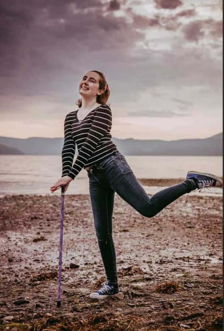 A young woman on a beach leaning on a stick with both hands and one foot playfully in the air.