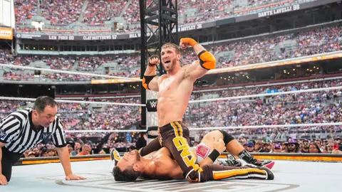 Getty Images Picture of a ring at a wrestling match. In the middle, Logan Paul has trapped his opponent on the floor and is astride him, and making a triumphant gesture to what looks like a massive arena audience. He is holding his arms aloft and sticking out his tongue. Next to them, a referee is on his hands and knees, monitoring the wrestling match.