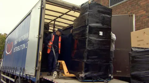 Steve Hubbard/BBC A pallet stacked with boxes covered in a black plastic wrap being unloaded from the back of a lorry. A man wearing a flat cap and high visibility jacket can be seen manoeuvring a hand-cranked forklift trolley under the pallet. Another man can just be seen behind the boxes wrapped in plastic. The cargo is stacked higher than both men and almost to the roof of the lorry. The side of the lorry can be seen on the left of the image, while in the foreground on the right is another stack of boxes wrapped in black plastic. 