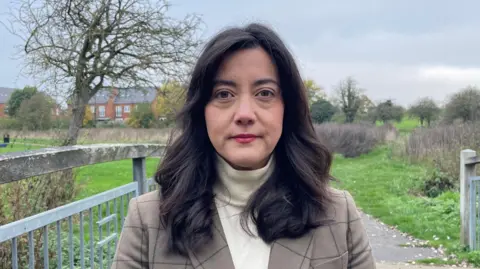 Ben Schofield/BBC Sarah Owen looking direct to camera. She is standing outside in an open space, with grass, trees and some housing in the background. To her left are fence-type railings. She is wearing a light brown jacket and cream high-necked top. She has shoulder length dark drown hair and brown eyes. 