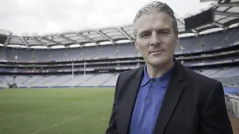 Jarlath Burns standing looking into the camera with a neutral expression on his face. He is wearing a suit jacket and blue polo shirt. He is standing in a large stadium with seats, a playing field and goal posts visible behind him. 