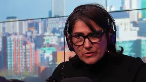 Dame Jasvinder Sanghera in a radio studio wearing glasses, a black top, and radio headphones