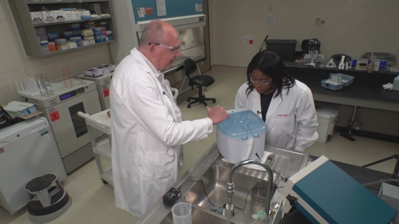 A man in a lab coat points at a small white, box line machine in a lab. A woman looks at where he is pointing.