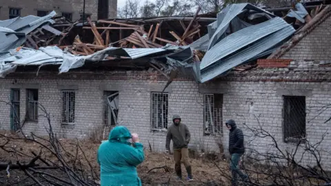 Reuters A woman and two men wearing coats with the hoods up stand in front of a one-story building with a destroyed roof and windows