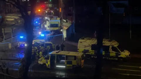 Police vans and a fire engine seen through the trees on a street by Chester station, with blue lights flashing and officers gathered on the road.