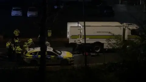 A bomb disposal van with the rear doors open next to a police vehicle with a group of police in high vis seen through the night sky. 