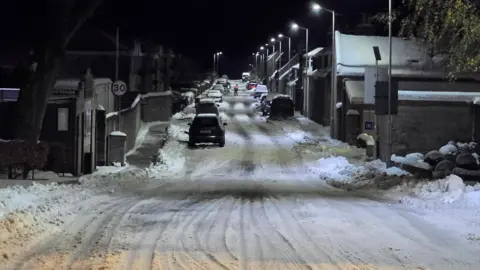 BBC Weather Watchers, Derek A snow covered road with cars and white streetlights extending into distance.