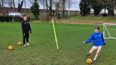 Andy Hughes Natasha Allen-Wyatt looks on as a young girl player kicks a ball on a football pitch