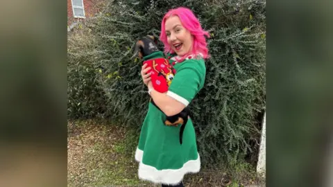 Zara Canfield A smiling woman stood in front of a tree while holding a dog. She has pink hair and is wearing a green dress with a white trim. The dog is also wearing a red jumper with an image of a cartoon dog on.