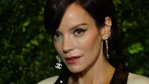 PA Media Lily Allen, pictured with black hair in an up do with a side fringe, winged eye liner and several diamond earrings