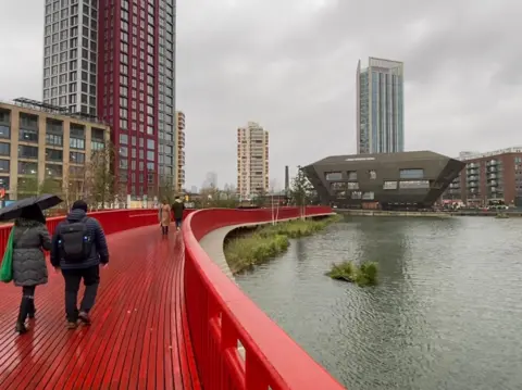 BBC Weather Watchers / Leroy D Grey skies in London, two people are seen sheltering under an umbrella as they walk over a red bridge