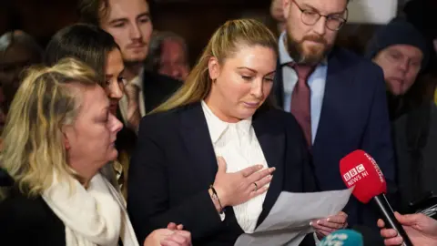 PA Media Nikita Hand, surrounded by two men and two women. She has blonde hair and is wearing a white top and black blazer. She has her hand on her chest and is looking at a piece of paper in her other hand. There is a red BBC News microphone held up in front of her.